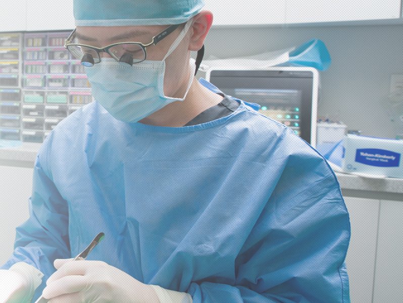 수술 모자·마스크·확대경을 쓴 파란 수술복 차림의 치과의사가 기구를 들고 수술 중(환자는 거의 안 보임)