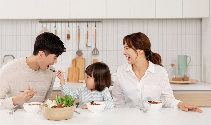 부엌 식탁에서 엄마·아빠·어린 딸이 웃으며 음식을 먹는 가족(아이가 아빠에게 먹여 줌)